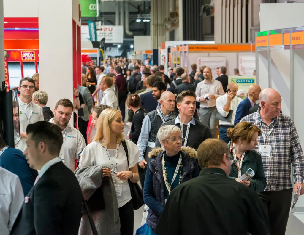 A crowded trade show floor at UK Construction Week with dozens of attendees wearing lanyards and badges networking among exhibition booths and displays with colorful signage.