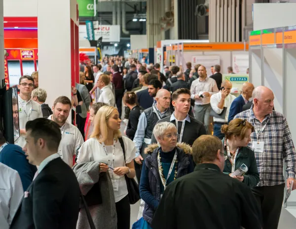A crowded trade show floor at UK Construction Week with dozens of attendees wearing lanyards and badges networking among exhibition booths and displays with colorful signage.