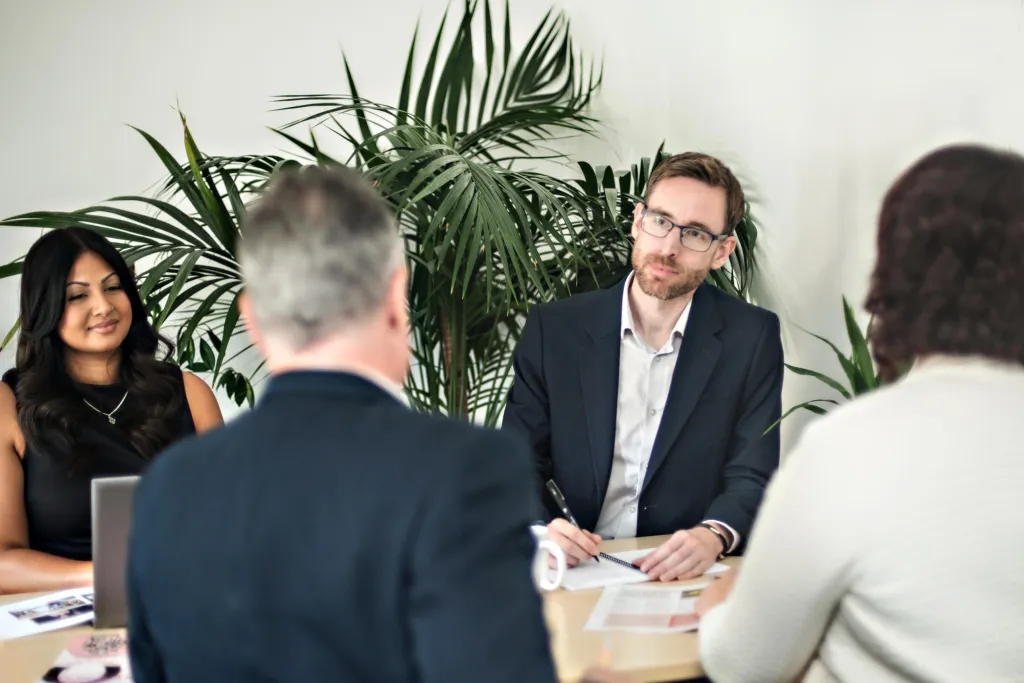A diverse team of four professionals sits around a conference table during a meeting in a bright office with a potted plant in the background. A man in glasses and dark blazer speaks with colleagues including a woman in black on the left and another woman on the right, while a fourth person with gray hair sits with their back to the camera.